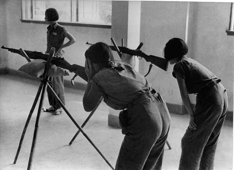 在一间室内射击场里，年轻的中国姑娘们在用三角架上的步枪练习瞄准。她们刻苦学习，争取提高射击技术。