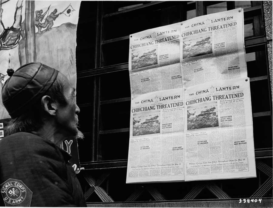 1945年5月8日。中国昆明，一位老者正在目光聚焦在在中国战区的官方报纸叶中国灯笼上，头条标题报道日本军队继续威胁进攻中国腹地。旁边的海报告诉人们德国已向盟军投降。 摄影师：约瑟夫